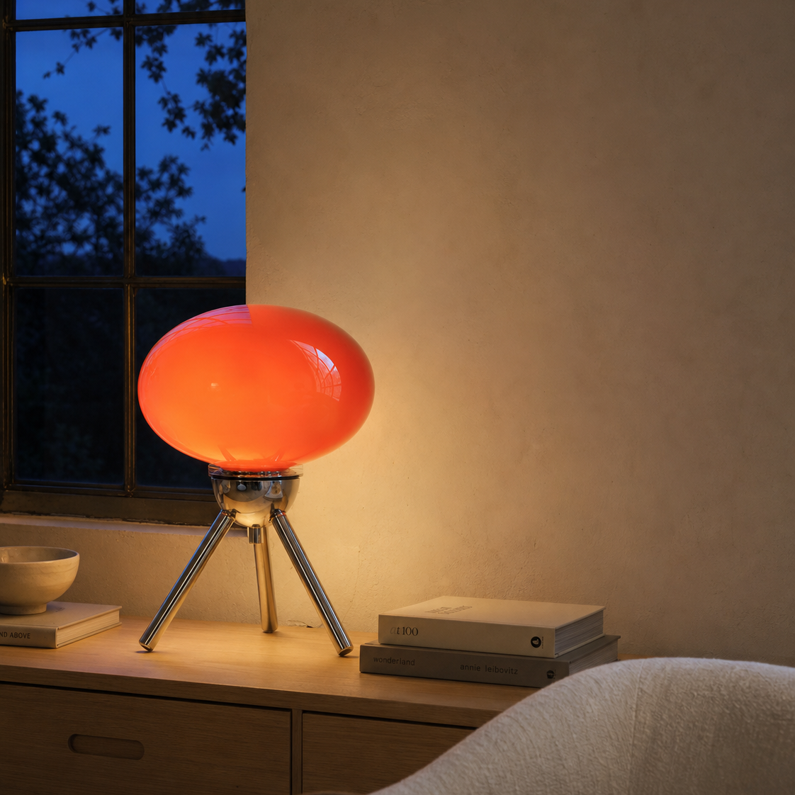 A modern table lamp with a glowing orange-red spherical shade on a silver tripod base, placed on a wooden console table in a warmly lit room.