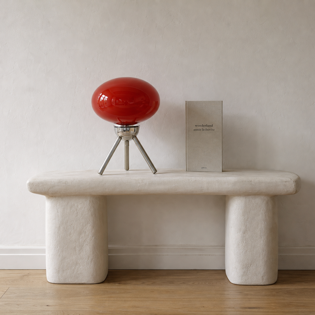 A modern sculptural bench made of textured off-white stone or concrete, styled with a glossy red spherical table lamp and a small book against a neutral beige wall and light wood floor.