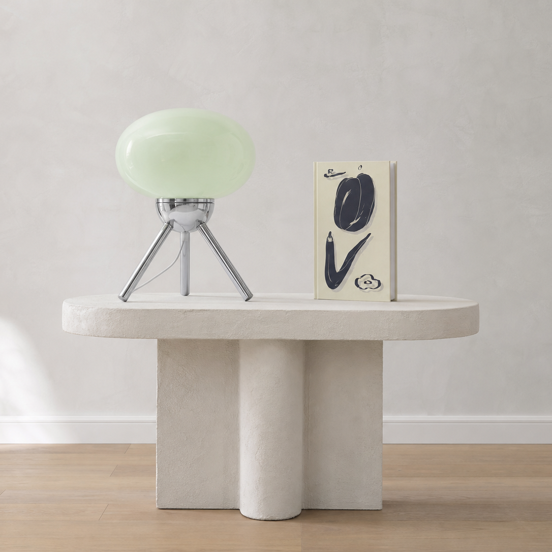 A modern sculptural side table in a light stone finish, styled with a pale green glass table lamp and a decorative book against a neutral textured wall.