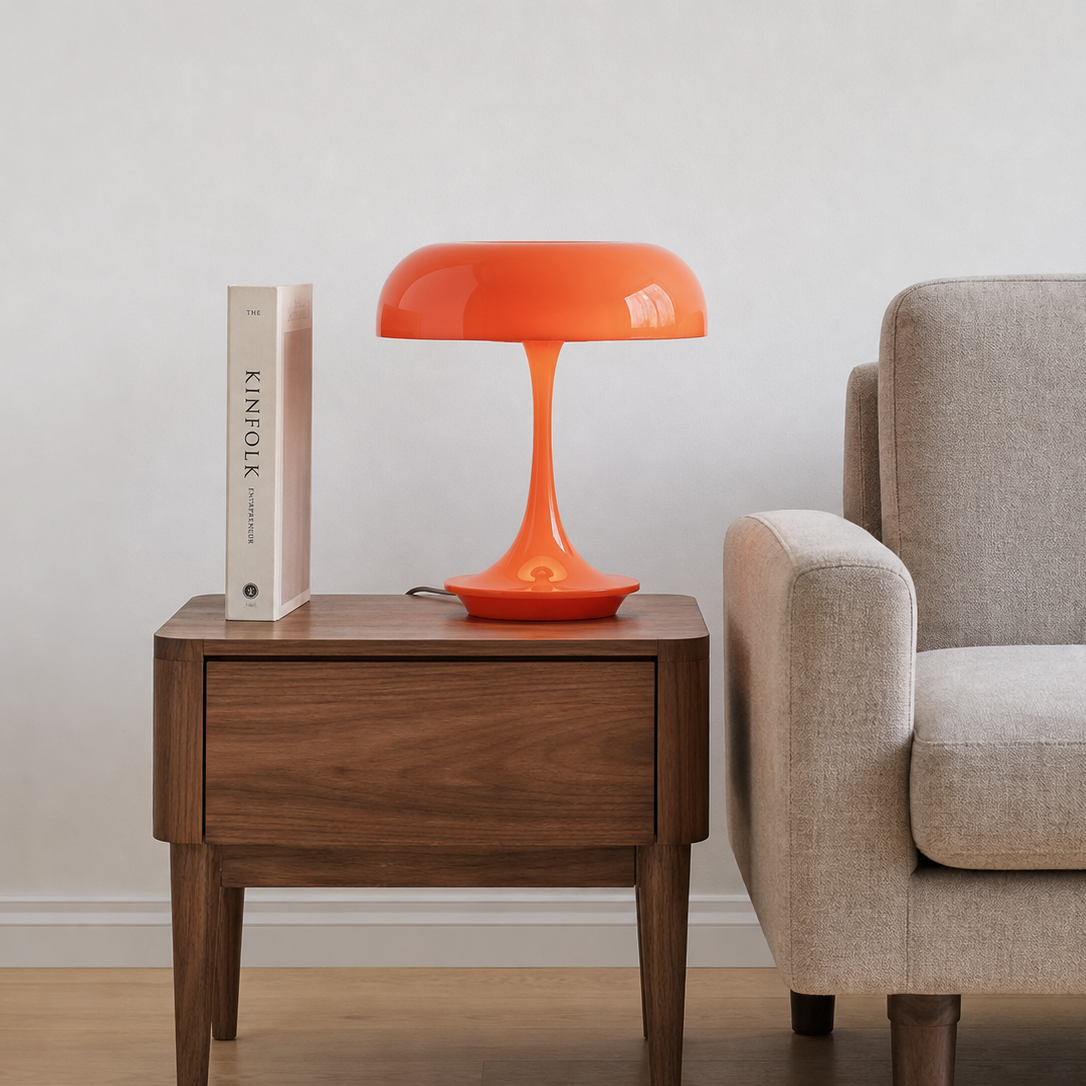 A bright orange mushroom-shaped table lamp sits on a dark wood side table next to a beige armchair, with a white book standing on top.