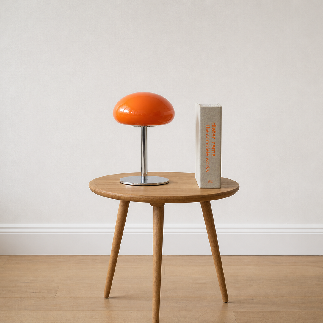 A round wooden side table with three tapered legs, styled with a bright orange mushroom-shaped table lamp and a tall gray book against a neutral off-white wall and light wood floor.