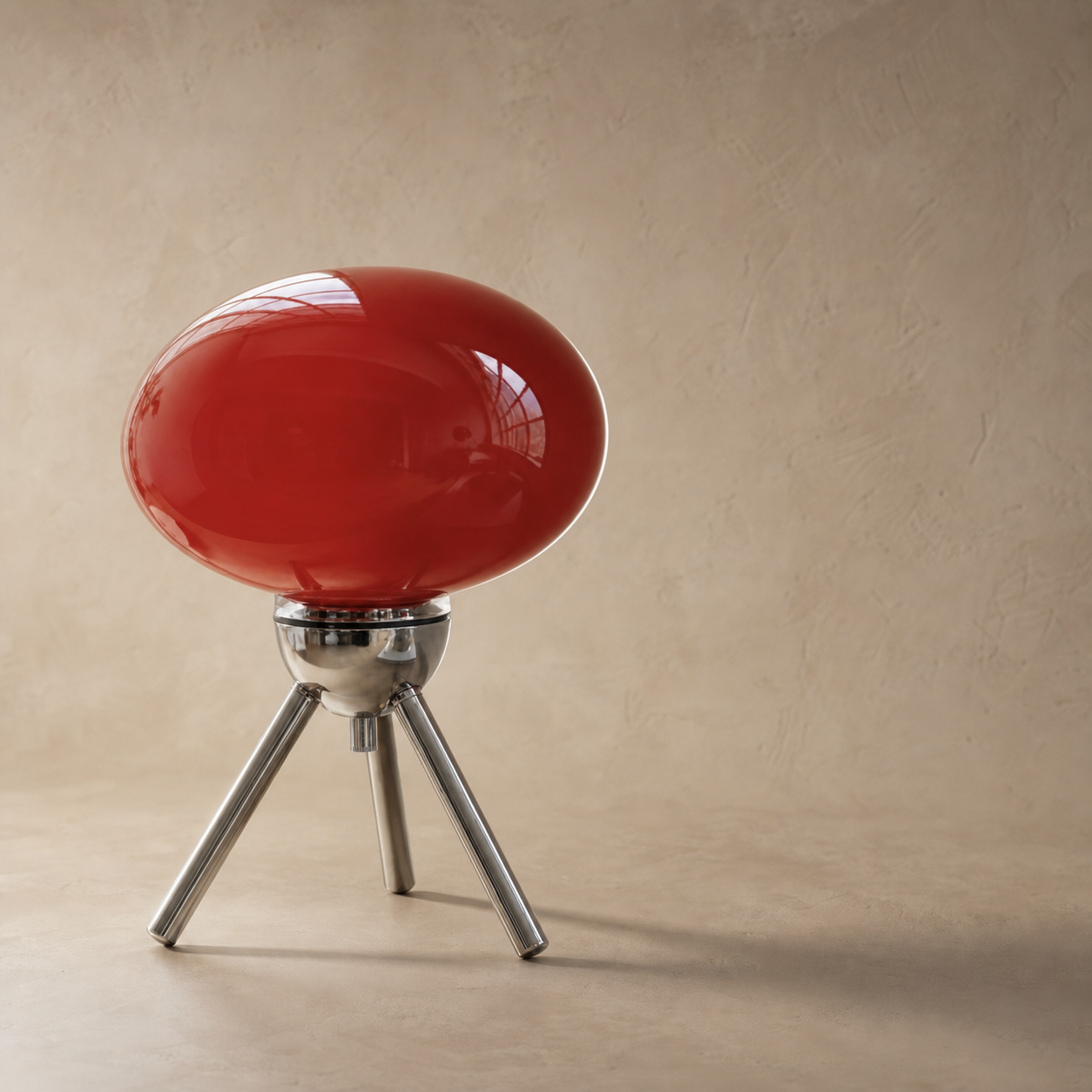 A modern table lamp featuring a glossy, egg-shaped red glass shade mounted on a polished silver metal tripod base, set against a warm beige textured wall.