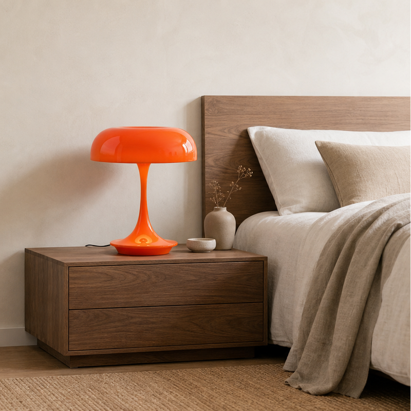 The Fungo mushroom table lamp in orange glowing on a dark wood nightstand beside a bed with neutral beige bedding