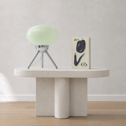 A modern sculptural side table in a light stone finish, styled with a pale green glass table lamp and a decorative book against a neutral textured wall.