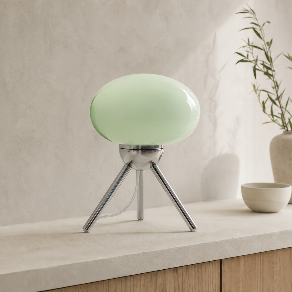 A modern table lamp with a glossy pale green oval shade on a three-legged silver metal tripod base, set on a light stone countertop against a textured beige wall.