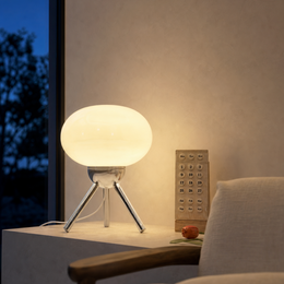 A modern table lamp with a glowing oval white shade and three silver legs sits on a white side table in a warmly lit room. A beige chair is partially visible in the foreground, and a small wooden object stands beside the lamp against a textured beige wall.