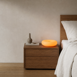 An orange glowing circular night light sits on a dark wood nightstand next to a bed with white linens in a minimalist bedroom setting.