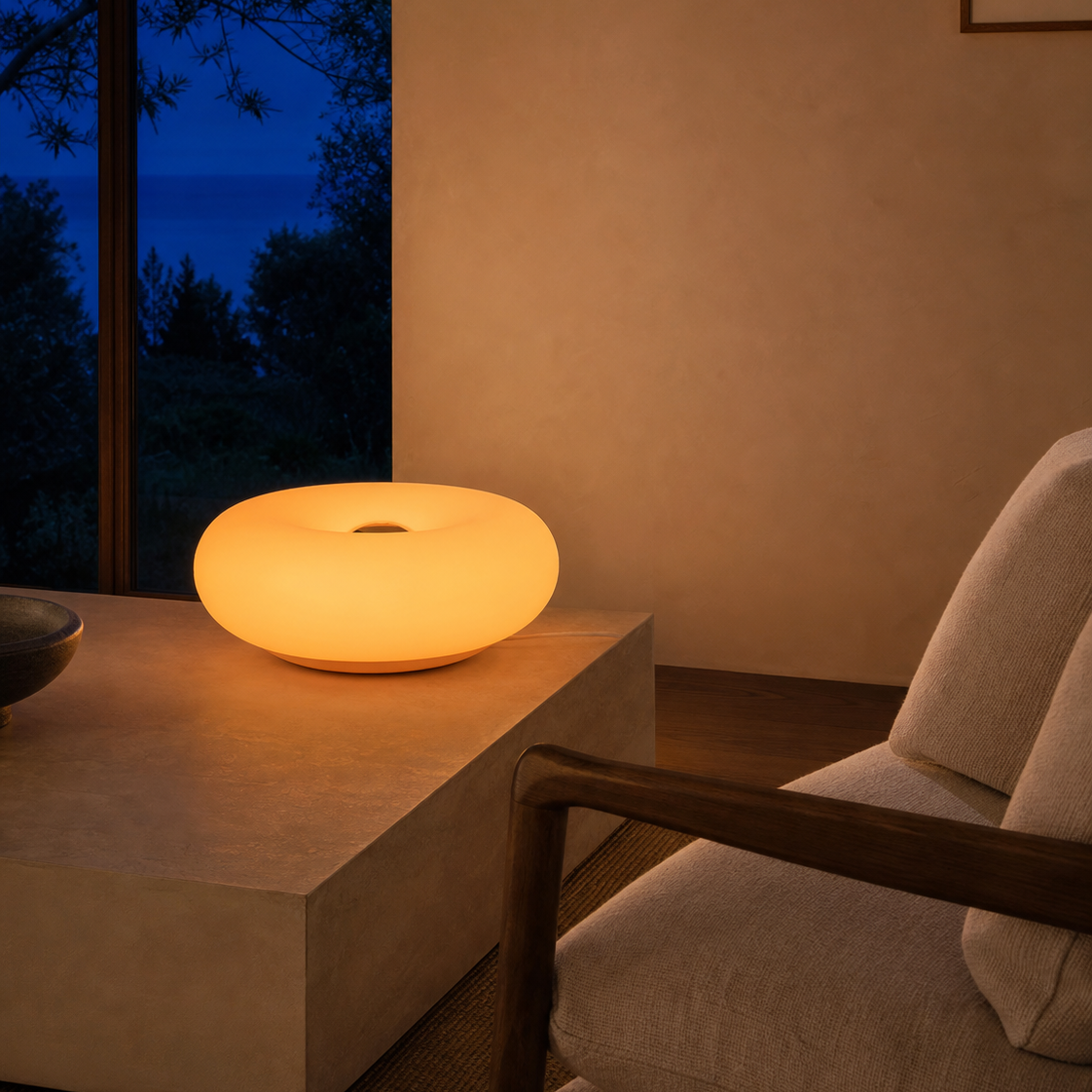 A glowing oval table lamp with a matte warm yellow-orange finish sits on a low concrete table in a minimalist room, next to a light beige upholstered armchair and a window showing a dark blue evening view.