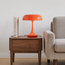 A bright orange mushroom-shaped table lamp sits on a dark wood side table next to a beige armchair, with a white book standing on top.