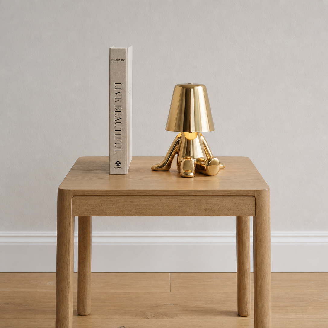 A modern brass table lamp with a tapered shade and three legs sits on a light wood side table next to a book titled 'LIVE BEAUTIFUL' against a neutral gray wall and light wood floor.