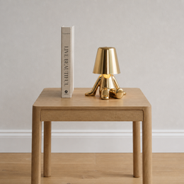 A modern brass table lamp with a tapered shade and three legs sits on a light wood side table next to a book titled 'LIVE BEAUTIFUL' against a neutral gray wall and light wood floor.