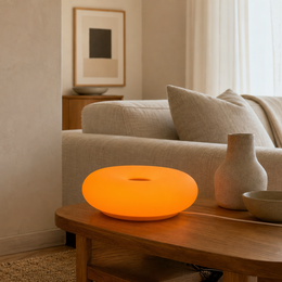A glowing orange oval smart lamp sits on a wooden coffee table in a modern, minimalist living room next to a beige sofa and neutral-toned decor.