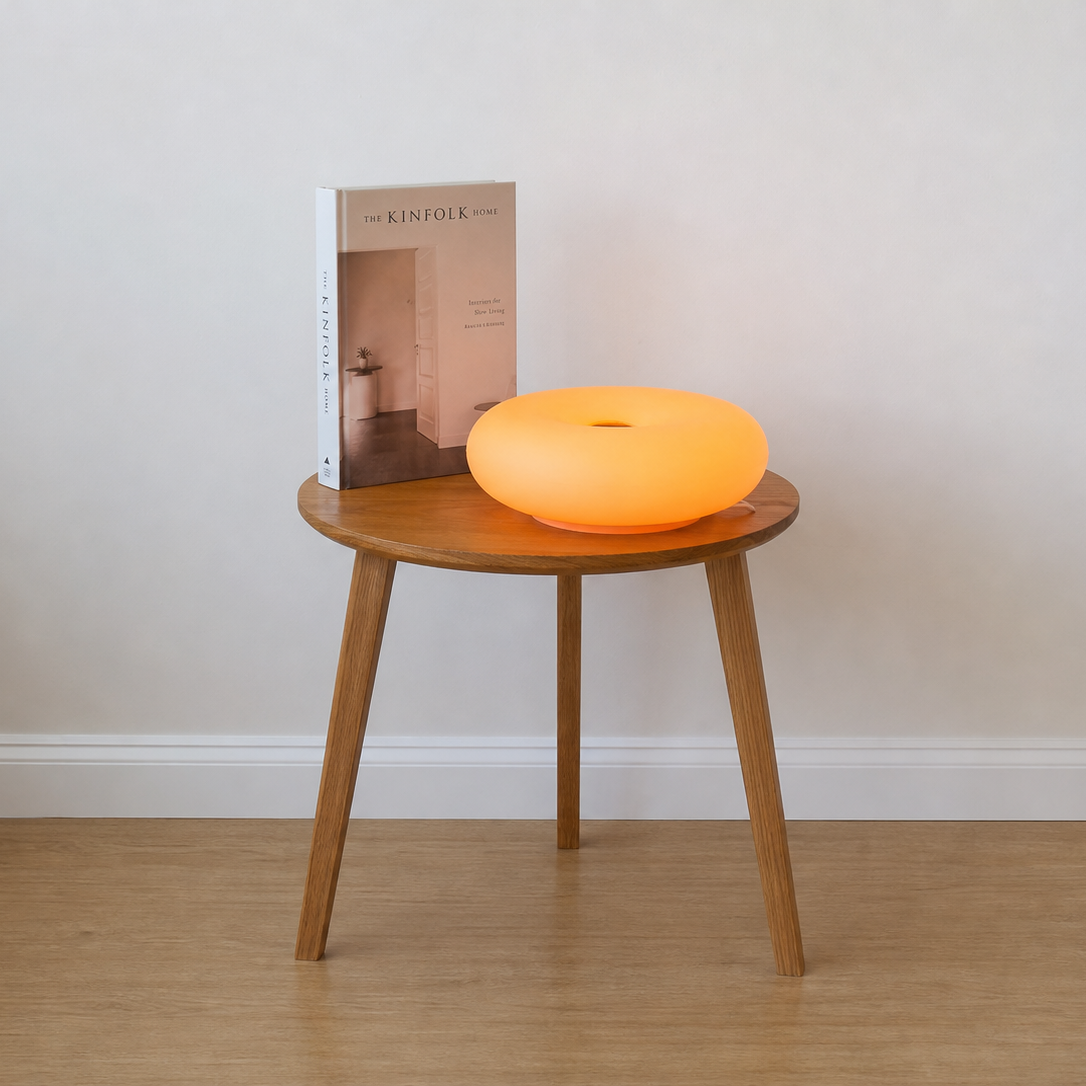An orange glowing donut-shaped lamp sits on a round wooden side table next to a book, set against a plain beige wall and light wood floor.