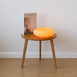 An orange glowing donut-shaped lamp sits on a round wooden side table next to a book, set against a plain beige wall and light wood floor.