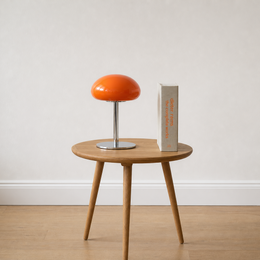 A round wooden side table with three tapered legs, styled with a bright orange mushroom-shaped table lamp and a tall gray book against a neutral off-white wall and light wood floor.