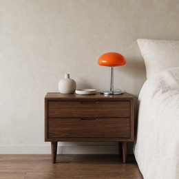 A mid-century modern two-drawer nightstand in dark walnut wood, styled next to a bed with white linens. On top of the nightstand sits a bright orange mushroom-shaped table lamp, a small beige ceramic vase, and a small white dish against a textured beige wall.