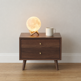 A mid-century modern wooden nightstand with two drawers and tapered legs, styled with a glowing moon lamp and a white ceramic mug against a neutral wall and light wood floor.