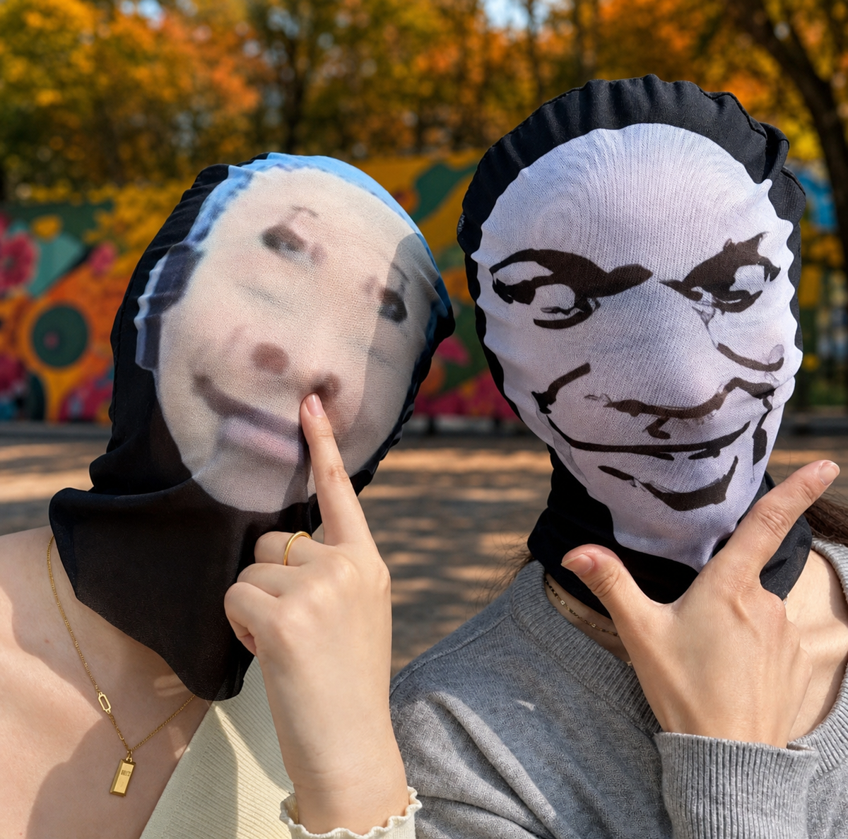Two people outdoors wearing printed face masks or hoodies over their heads, with one person pointing at the printed face on the left and the other wearing a white printed face on the right. Trees and a colorful mural are visible in the background.