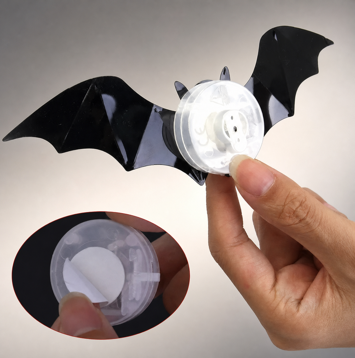 A black bat-shaped adhesive sticker held by a hand, with a circular inset showing the clear adhesive backing and internal mechanism.