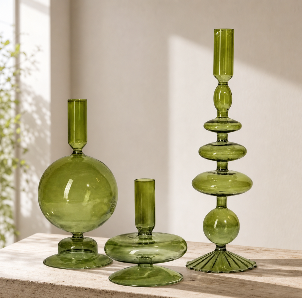 Three green glass decorative vessels on a light wood surface against a warm beige wall, including a tall fluted pedestal candle holder, a round bulb vase, and a short matching candle holder.