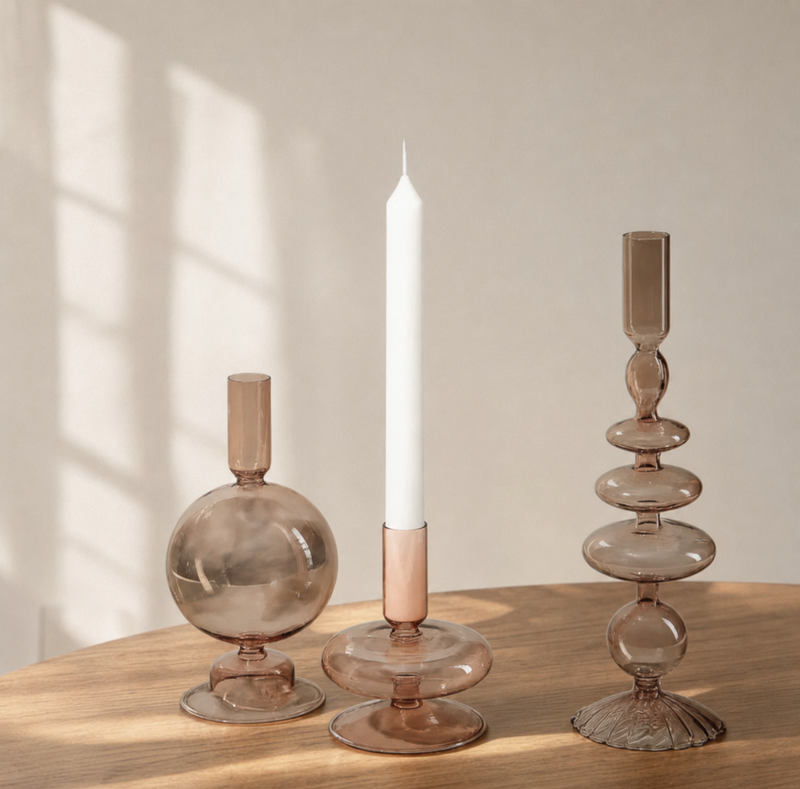 Three amber-tinted glass candle holders on a wooden table, with a white taper candle standing in the center holder. Soft window shadows fall on the beige wall behind.