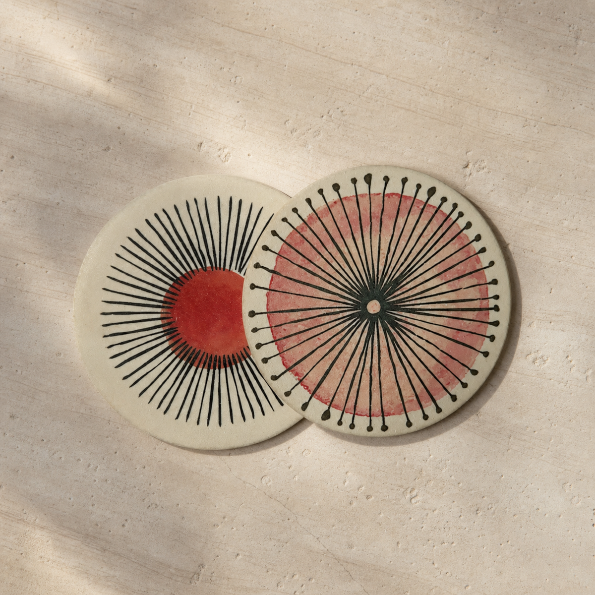Two round ceramic coasters with mid-century modern sunburst designs, displayed on a textured beige stone surface with soft leaf shadows.