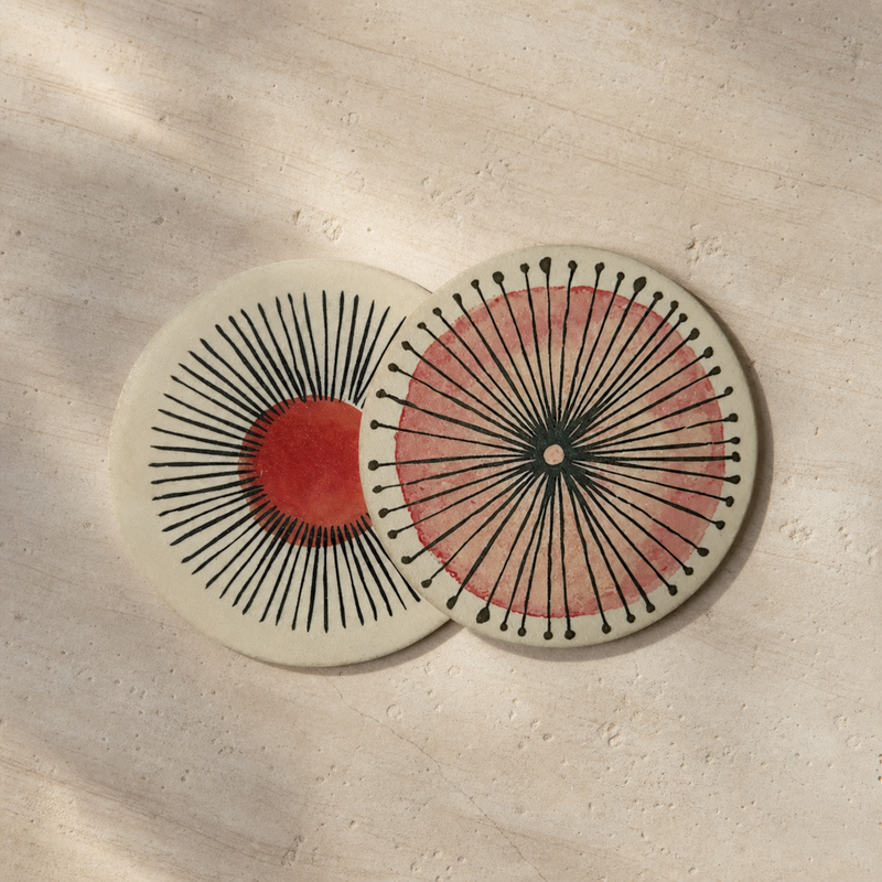 Two round ceramic coasters with mid-century modern sunburst designs, displayed on a textured beige stone surface with soft leaf shadows.