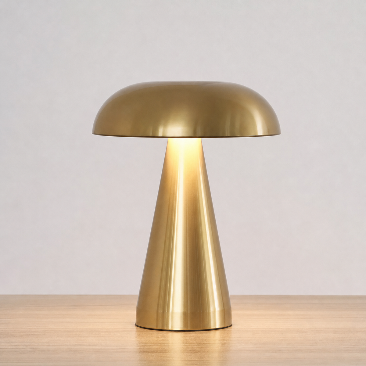A modern mushroom-shaped table lamp with a brushed brass finish, illuminated from within, sitting on a light wood surface against a neutral light gray background.