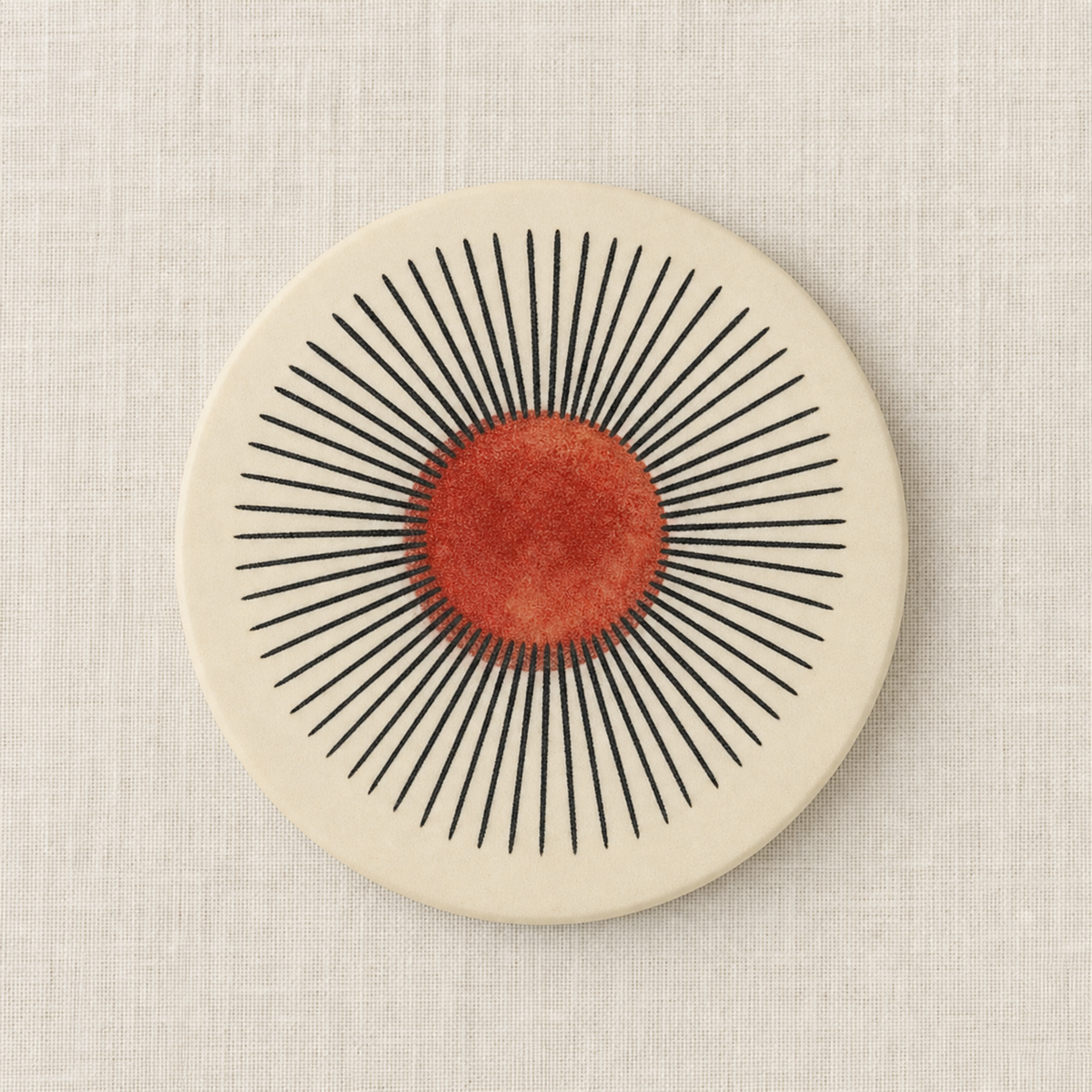 A round ceramic plate or coaster with a minimalist mid-century modern design, featuring a textured terracotta orange-red sunburst pattern on a warm cream background, set against a neutral linen backdrop.