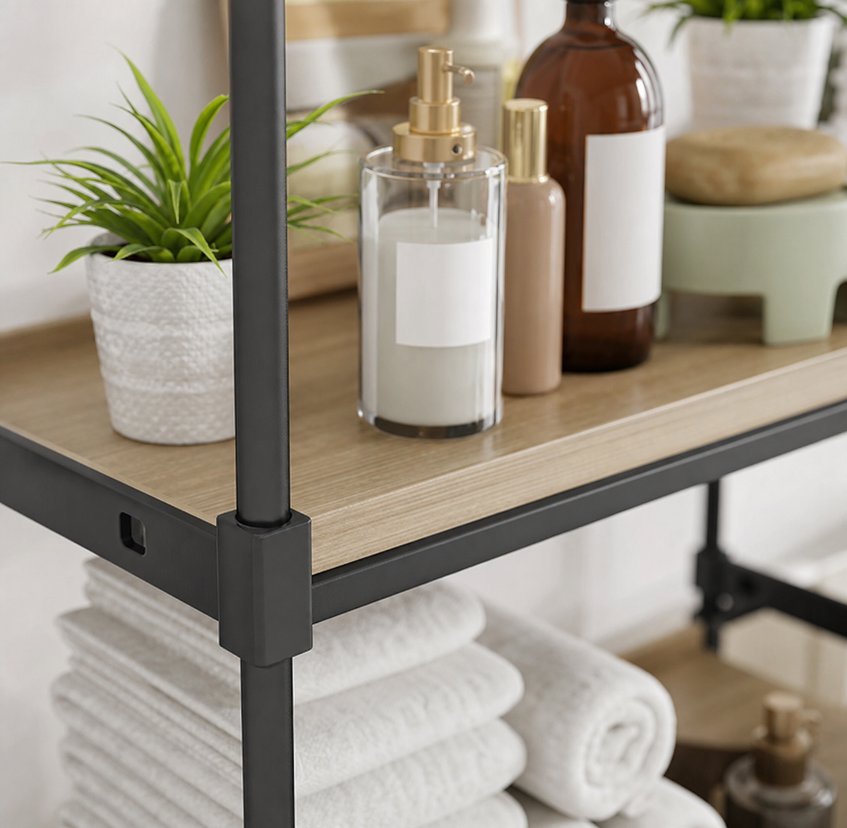 A modern industrial shelving unit with a light wood grain top and black metal frame, styled with a small potted plant, a glass candle jar with a gold pump, an amber bottle, and folded white towels on the lower shelf.