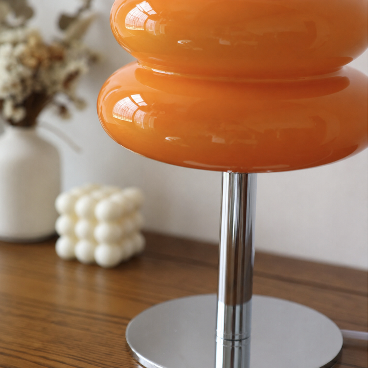 An orange glossy table lamp with a tiered mushroom-shaped shade and a chrome metal base, shown on a wooden table with a blurred background.