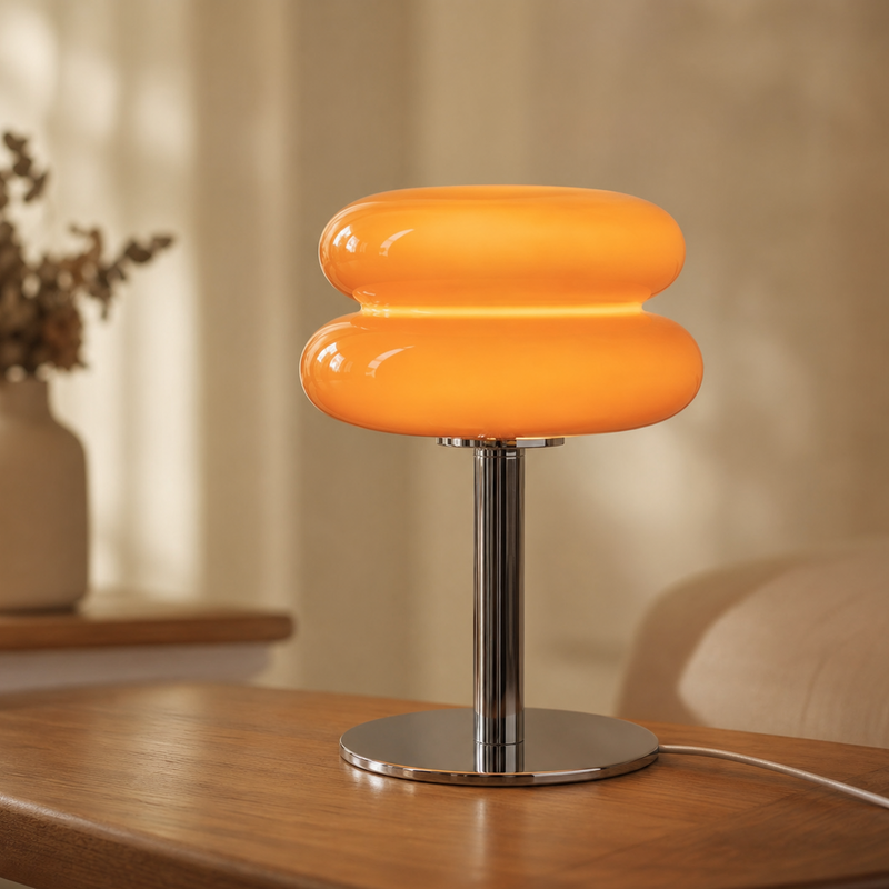 A modern table lamp with a glossy orange, two-bulb-shaped shade on a polished chrome stand, placed on a wooden table in a warm, softly lit room.