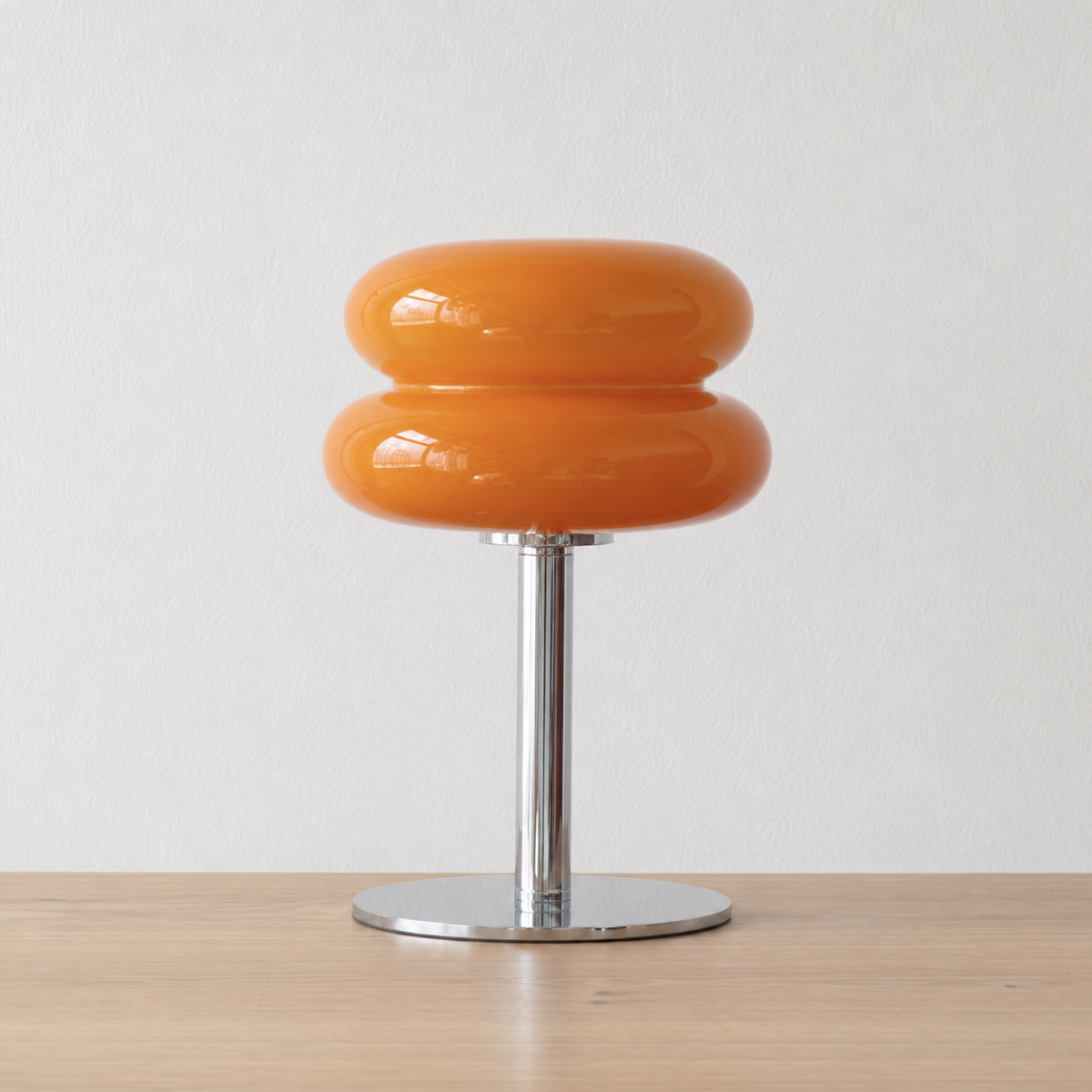 An orange glossy two-ring table lamp with a chrome metal stem and circular base, set on a light wood surface against a white wall.