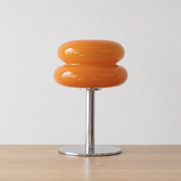 An orange glossy two-ring table lamp with a chrome metal stem and circular base, set on a light wood surface against a white wall.