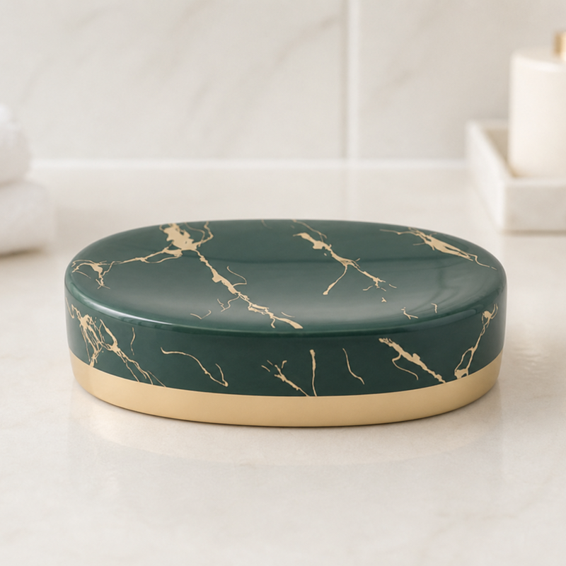 An oval soap dish with a glossy dark green marble pattern and gold metallic trim, sitting on a white marble countertop with a folded towel and bathroom accessories blurred in the background.
