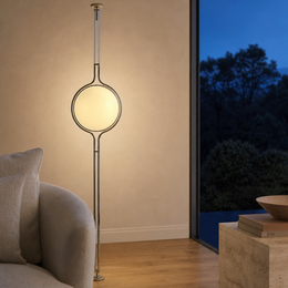 A modern floor lamp with a glowing oval shade and a slim metallic frame, standing in a warm-toned room next to a window looking out onto a dark evening sky.