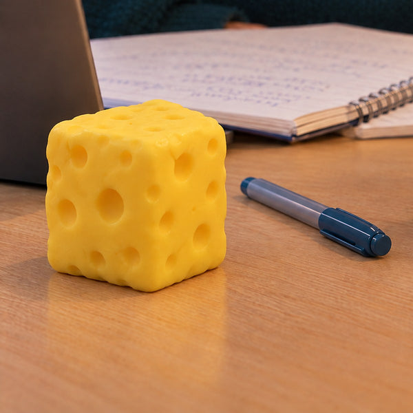 A yellow cheese squishy cube on a wooden desk next to a pen, a notebook and a laptop (styled illustration)