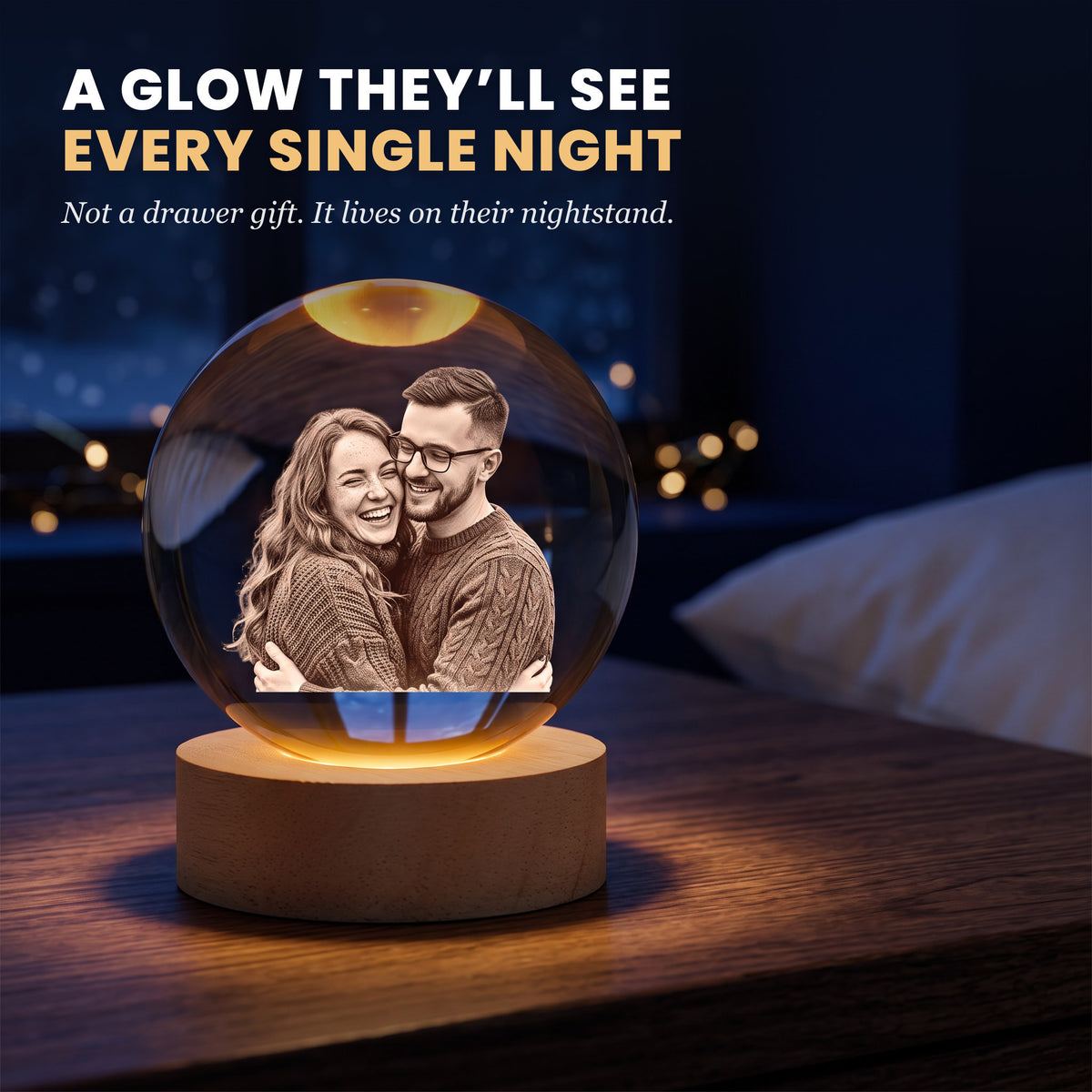 Crystal ball night light glowing warm on a nightstand at night, a laughing couple engraved inside, snowy window behind